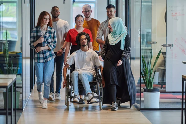 現代のスタートアップのガラス張りのオフィスで廊下を歩く若いビジネスの人々の多様なグループは 車椅子やヒジャブを着用している女性を含み ダイナミックなミックスを示しています — ストック写真