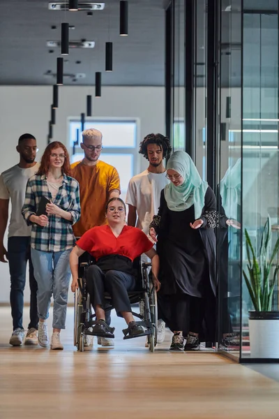 stock image A diverse group of young business people walking a corridor in the glass-enclosed office of a modern startup, including a person in a wheelchair and a woman wearing a hijab, showing a dynamic mix of