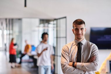 Genç bir iş adamı modern bir ofis koridorunda kollarını kavuşturarak duruyor. Kendine güven ve bir amaç duygusu yayıyor. Dinamik ve ilham verici bir varlığı somutlaştırıyor.