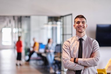 Genç bir iş adamı modern bir ofis koridorunda kollarını kavuşturarak duruyor. Kendine güven ve bir amaç duygusu yayıyor. Dinamik ve ilham verici bir varlığı somutlaştırıyor.