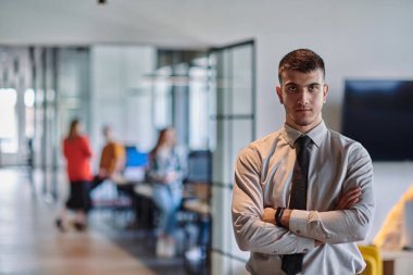 Genç bir iş adamı modern bir ofis koridorunda kollarını kavuşturarak duruyor. Kendine güven ve bir amaç duygusu yayıyor. Dinamik ve ilham verici bir varlığı somutlaştırıyor.