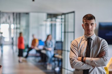 Genç bir iş adamı modern bir ofis koridorunda kollarını kavuşturarak duruyor. Kendine güven ve bir amaç duygusu yayıyor. Dinamik ve ilham verici bir varlığı somutlaştırıyor.