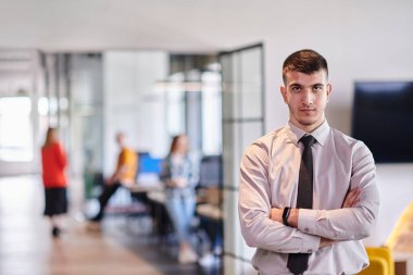 Genç bir iş adamı modern bir ofis koridorunda kollarını kavuşturarak duruyor. Kendine güven ve bir amaç duygusu yayıyor. Dinamik ve ilham verici bir varlığı somutlaştırıyor.