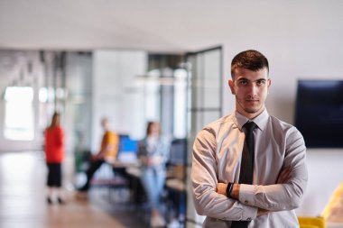 Genç bir iş adamı modern bir ofis koridorunda kollarını kavuşturarak duruyor. Kendine güven ve bir amaç duygusu yayıyor. Dinamik ve ilham verici bir varlığı somutlaştırıyor.