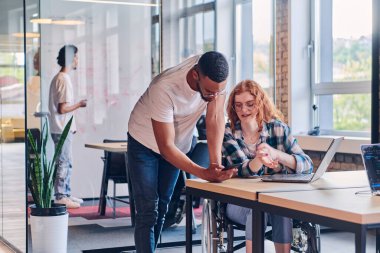 Tekerlekli sandalyedeki Afrikalı Amerikalı bir işadamı da dahil olmak üzere bir iş grubu, modern bir cam ofis içinde, bilgisayar ve dizüstü bilgisayarın etrafında aktif olarak iş birliği yaparak çeşitli çözümler üretiyor.