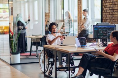 Aralarında tekerlekli sandalyedeki Afrikalı Amerikalı bir işadamının da bulunduğu genç bir iş adamı, modern bir cam ofis içinde, bir bilgisayar ve dizüstü bilgisayarın etrafında aktif olarak iş birliği yaparak,