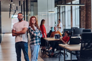 Genç bir Afrikalı Amerikalı işadamı ve turuncu saçlı modern bir iş kadını kollarını kavuşturmuş, çağdaş ofis ortamında güven ve birlik saçan, dinamiği simgeleyen