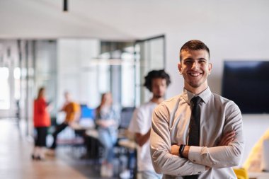 Genç bir iş adamı modern bir ofis koridorunda kollarını kavuşturarak duruyor. Kendine güven ve bir amaç duygusu yayıyor. Dinamik ve ilham verici bir varlığı somutlaştırıyor.