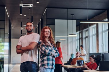 Genç bir Afrikalı Amerikalı işadamı ve turuncu saçlı modern bir iş kadını kollarını kavuşturmuş, çağdaş ofis ortamında güven ve birlik saçan, dinamiği simgeleyen