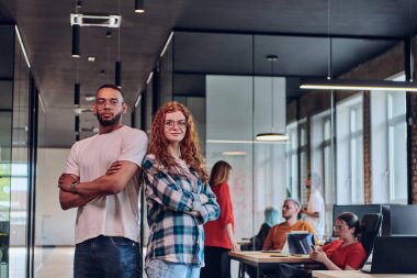 Genç bir Afrikalı Amerikalı işadamı ve turuncu saçlı modern bir iş kadını kollarını kavuşturmuş, çağdaş ofis ortamında güven ve birlik saçan, dinamiği simgeleyen