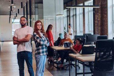 Genç bir Afrikalı Amerikalı işadamı ve turuncu saçlı modern bir iş kadını kollarını kavuşturmuş, çağdaş ofis ortamında güven ve birlik saçan, dinamiği simgeleyen