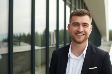 Pürüzsüz siyah takım elbiseli bir CEO modern bir şirket binasının girişinde duruyor ve hareketli şehir ortamında iş gününün başlamasını bekliyor.