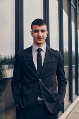 Pürüzsüz siyah takım elbiseli bir CEO modern bir şirket binasının girişinde duruyor ve hareketli şehir ortamında iş gününün başlamasını bekliyor.