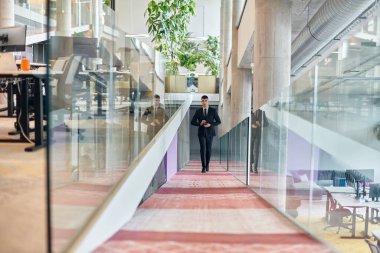 Gösterişli bir şirket ortamında, genç ve karizmatik yönetmen koridorda güven içinde yürüyor, dinamik liderliğini ve şirket içindeki başarısının devamını simgeliyor.