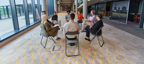 A diverse group of young business entrepreneurs gathered in a circle for a meeting, discussing corporate challenges and innovative solutions within the modern confines of a large corporation.