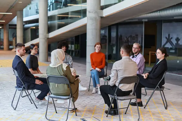 A diverse group of young business entrepreneurs gathered in a circle for a meeting, discussing corporate challenges and innovative solutions within the modern confines of a large corporation.