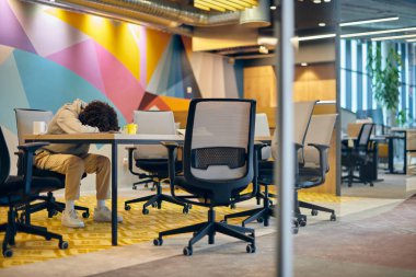 Modern, geniş bir ofiste, yorgun bir Afrikalı Amerikalı adam masa başında uzanmış, çağdaş kurumsal yaşamın zorluklarını ve iş-yaşam dengesinin önemini yansıtıyor.
