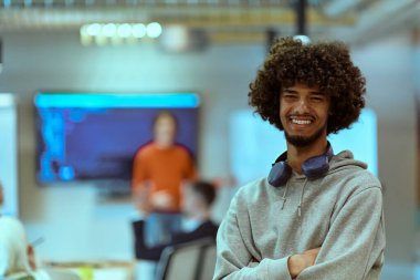 Modern bir ofis ortamında, kulaklıklı bir Afrikalı Amerikalı genç girişimci işe koyulurken, arka planda, kendini işine adamış meslektaşları takım çalışması ve işbirliğini örnek gösteriyorlar.