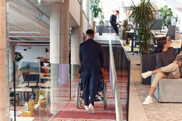 Директор компании помогает своей коллеге по бизнесу в инвалидной коляске, помогая ей ориентироваться в их стартап-офисе, где они работают вместе со своими разнообразными коллегами, подчеркивая инклюзивность и