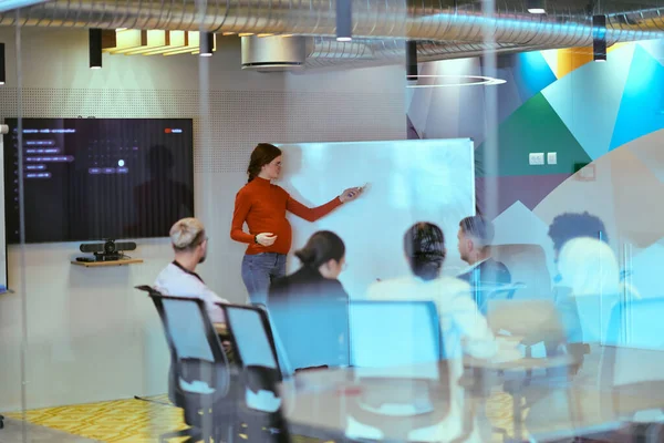 A pregnant business woman with orange hair confidently presents her business plan to colleagues in a modern glass office, embodying entrepreneurship and innovation.