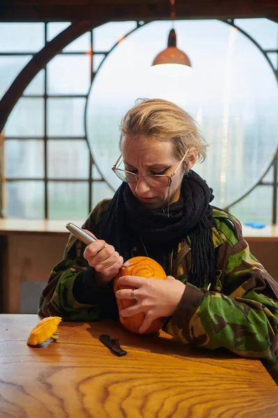 軍服の現代のブロンドの女性はハロウィーンの夜のナイフで不気味なカボチャを彫っています — ストック写真