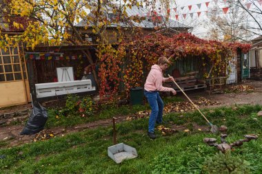 Bir adam, bahçeyi özenle eski, kuru yaprakları toplayarak, açık hava bakımının ve mevsimsel düzenliliğin resimli bir sahnesini oluşturarak koruyor.