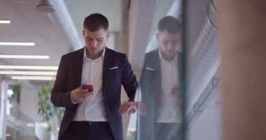 Modern bir şirket koridorunda meşgul bir işadamı, yoğun iş programını yönetmek ve hızlı iş dünyasıyla bağlantıda kalmak için akıllı telefon kullanarak birden fazla iş yapma becerisini sergiliyor.