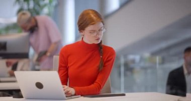 Modern bir ofiste, turuncu saçlı profesyonel bir iş kadını dizüstü bilgisayarında oturarak, çağdaş çalışma alanındaki yenilikleri ve verimliliği özetliyor.. 