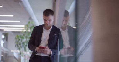 Modern bir şirket koridorunda meşgul bir işadamı, yoğun iş programını yönetmek ve hızlı iş dünyasıyla bağlantıda kalmak için akıllı telefon kullanarak birden fazla iş yapma becerisini sergiliyor..