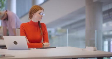 Modern bir ofiste, turuncu saçlı profesyonel bir iş kadını dizüstü bilgisayarında oturarak, çağdaş çalışma alanındaki yenilikleri ve verimliliği özetliyor.