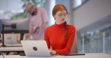 Modern bir ofiste, turuncu saçlı profesyonel bir iş kadını dizüstü bilgisayarında oturarak, çağdaş çalışma alanındaki yenilikleri ve verimliliği özetliyor.