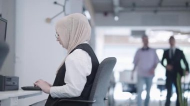 Modern bir ofiste tesettürlü bir iş kadını çalışırken, çeşitli meslektaşları yanından geçerken, özveri, profesyonellik ve çok kültürlü takım çalışmalarını örnek olarak görülüyor.