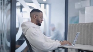 Afrika kökenli Amerikalı bir adam modern ofis ortamında özenle dizüstü bilgisayarında çalışırken, çeşitli meslektaşları onun etrafında hareket ediyor ve odaklanmış ve üretken bir çalışma alanı çiziyor.. 