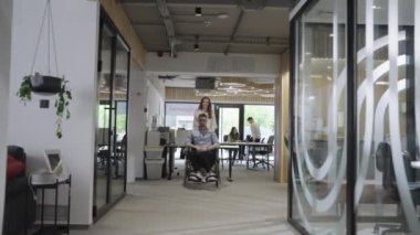 Genç iş arkadaşları, işbirlikçi iş arkadaşları, aralarında tekerlekli sandalyedeki bir kişinin de bulunduğu, modern cam ofis koridorunun yanından geçerek, çeşitliliği, takım çalışmasını ve güçlendirmeyi resmediyorlar.
