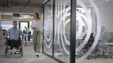 Genç iş arkadaşları, işbirlikçi iş arkadaşları, aralarında tekerlekli sandalyedeki bir kişinin de bulunduğu, modern cam ofis koridorunun yanından geçerek, çeşitliliği, takım çalışmasını ve güçlendirmeyi resmediyorlar.