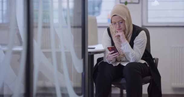 一位身披头巾的女商人在一个现代的办公室里 用她的智能手机在一个现代的工作空间里休息一下 突出强调了多样性 科技的平衡和创新 — 图库视频影像