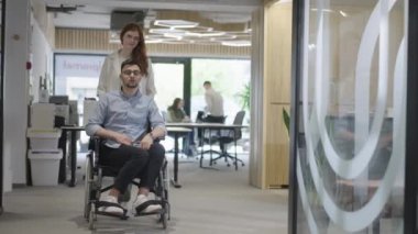 Genç iş arkadaşları, işbirlikçi iş arkadaşları, aralarında tekerlekli sandalyedeki bir kişinin de bulunduğu, modern cam ofis koridorunun yanından geçerek, çeşitliliği, takım çalışmasını ve güçlendirmeyi resmediyorlar.