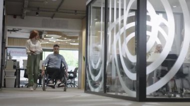 Genç iş arkadaşları, işbirlikçi iş arkadaşları, aralarında tekerlekli sandalyedeki bir kişinin de bulunduğu, modern cam ofis koridorunun yanından geçerek, çeşitliliği, takım çalışmasını ve güçlendirmeyi resmediyorlar.
