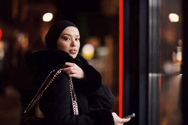 Soğuk bir kış gecesinde şehir caddesinde yürüyen Müslüman bir kadın arka planda Bokeh şehir ışıklarıyla tesettüre girmiş.