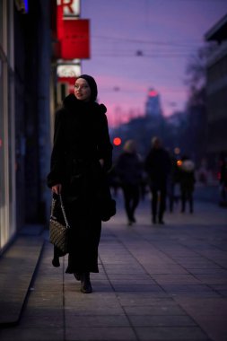 Soğuk bir kış gecesinde şehir caddesinde yürüyen Müslüman bir kadın arka planda Bokeh şehir ışıklarıyla tesettüre girmiş.