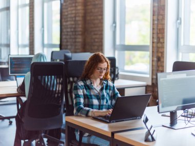 Canlı turuncu saçlı, genç ve başarılı bir iş kadını modern bir ofis içinde odaklanmış bir şekilde dizüstü bilgisayarını kullanarak özgüvenini ve dinamik yaklaşımını sergiliyor.