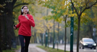 Sağlıklı yaşam tarzı, sabah koşusu. Genç ve güzel bir kadın sonbahar parkında koşuyor ve akıllı telefonuyla kulaklıkla müzik dinliyor. Kırmızı bir spor gömlek giyiyor.. 