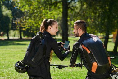 Bisikletlerle donatılmış ve yolculuklarını koordine etmeye odaklanmış tatlı bir çift, parkta manzaralı rotalar planlarken GPS mobil ve saatlerini kontrol ediyor, kusursuz bir şekilde harmanlama teknolojisi ve