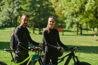 Profesyonel bisiklet kıyafetleriyle süslenmiş mutlu bir çift, parkın içinde romantik bir bisiklet sürmekten hoşlanıyor, modern doğal cazibelerle çevrili, sevgi ve mutluluk saçıyor.. 