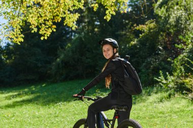 Güneşli bir günün ışıltılı kucaklamasında profesyonel bisiklet kıyafetleriyle süslenmiş neşeli bir kız, parkta bisikletle dolaşırken saf mutluluk ve canlılık bulur, bulaşıcı gülüşü