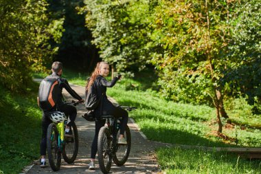 Profesyonel bisiklet kıyafetleriyle süslenmiş mutlu bir çift, parkın içinde romantik bir bisiklet sürmekten hoşlanıyor, modern doğal cazibelerle çevrili, sevgi ve mutluluk saçıyor.. 