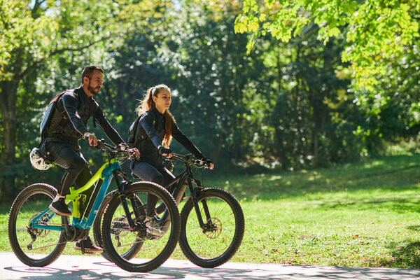 Блаженная пара, украшенная профессиональным велосипедным снаряжением, наслаждается романтической поездкой на велосипеде через парк, окруженный современными природными достопримечательностями, излучающими любовь и счастье. 