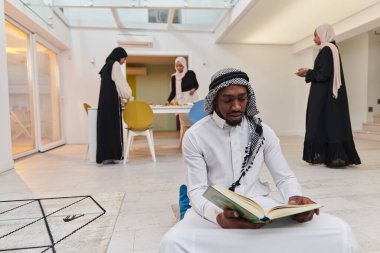 Kutsal Ramazan ayında, Kuran okumaya dalmış Afrikalı bir Müslüman adam üç tesettürlü kadının iftar için yemek hazırlayıp servis ettiği bir zemin ile çevrilidir.