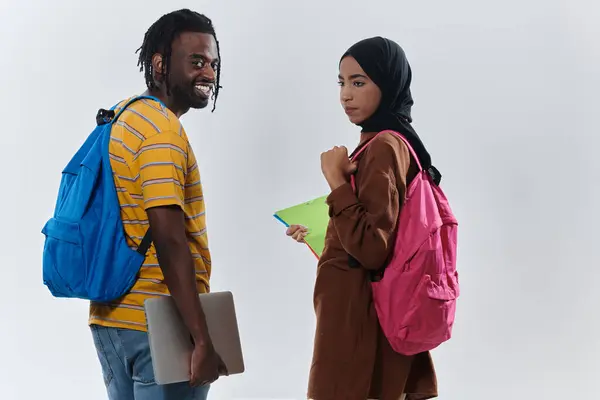 African American student collaborates with his Muslim colleague, who diligently works on her laptop, symbolizing a blend of diversity, modern learning, and cooperative spirit against a serene white