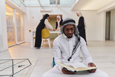 Kutsal Ramazan ayında, Kuran okumaya dalmış Afrikalı bir Müslüman adam üç tesettürlü kadının iftar için yemek hazırlayıp servis ettiği bir zemin ile çevrilidir.